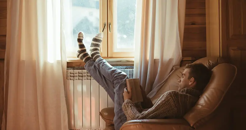 Young man in warm sweater reading book while relaxing on armchair by the window and electric radiator inside cozy log cabin